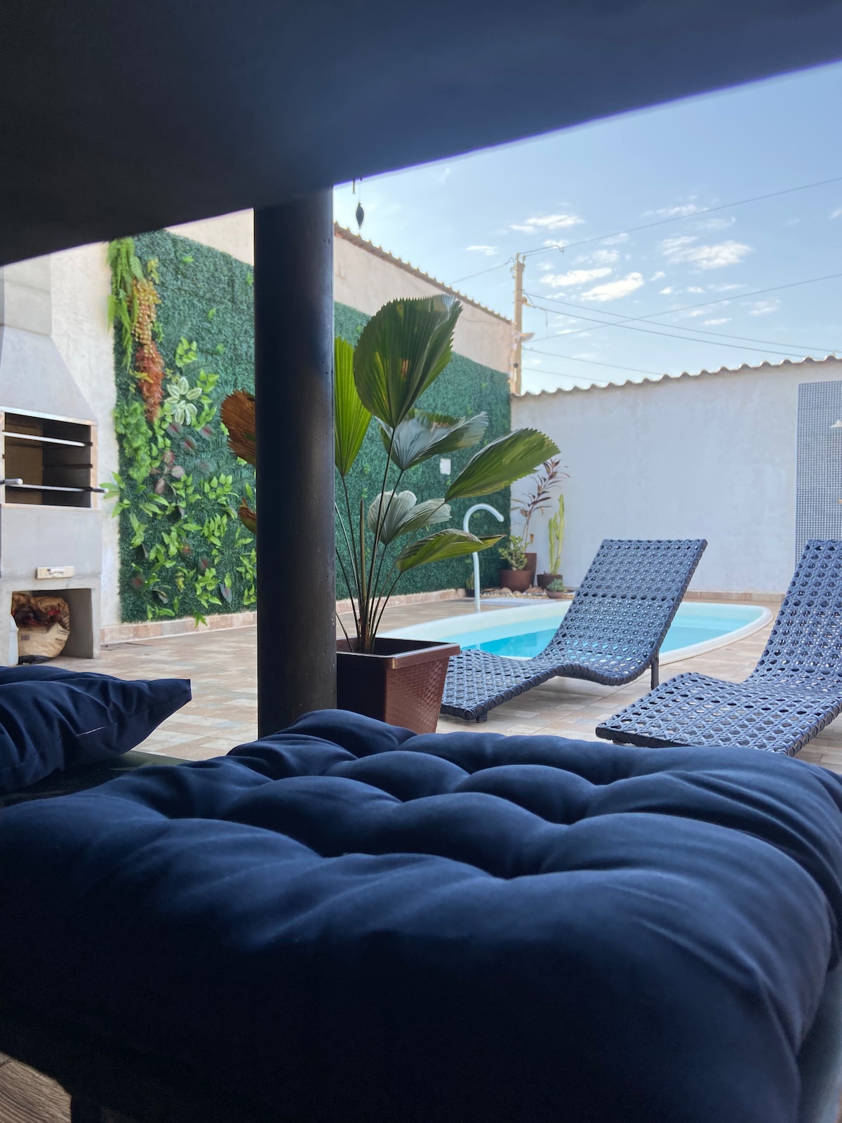 A clear view of a tranquil outdoor area is presented, featuring a pool surrounded by two lounge chairs. A vibrant green vertical garden adds a natural touch, while a barbecue area is visible in the background. Soft cushions lie on a nearby seating area.