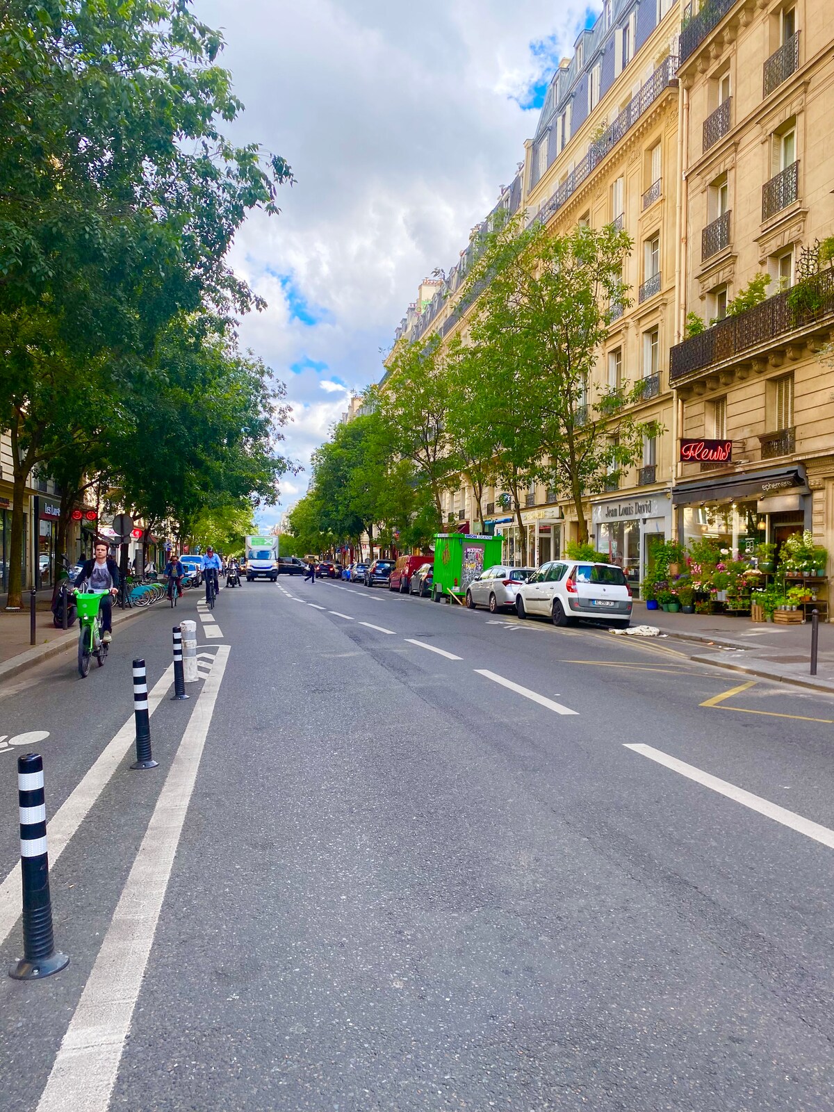 A wide street in Paris is lined with trees and buildings featuring shopfronts and parked vehicles. Bicycles can be seen on the bike lane, and pedestrians walk along the sidewalk, illustrating the vibrant urban atmosphere.