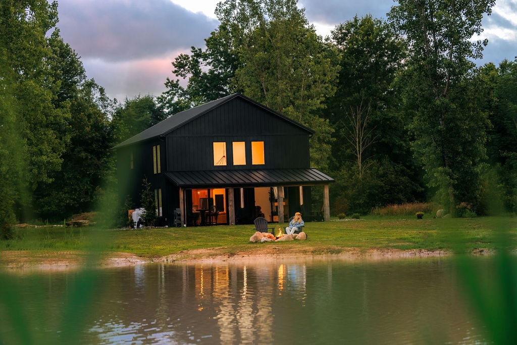 A two-story black cabin is situated beside a tranquil pond, with warm light illuminating its windows. Lush green trees surround the property, creating a serene atmosphere. In the foreground, individuals are observed near the water's edge, enjoying the natural setting.