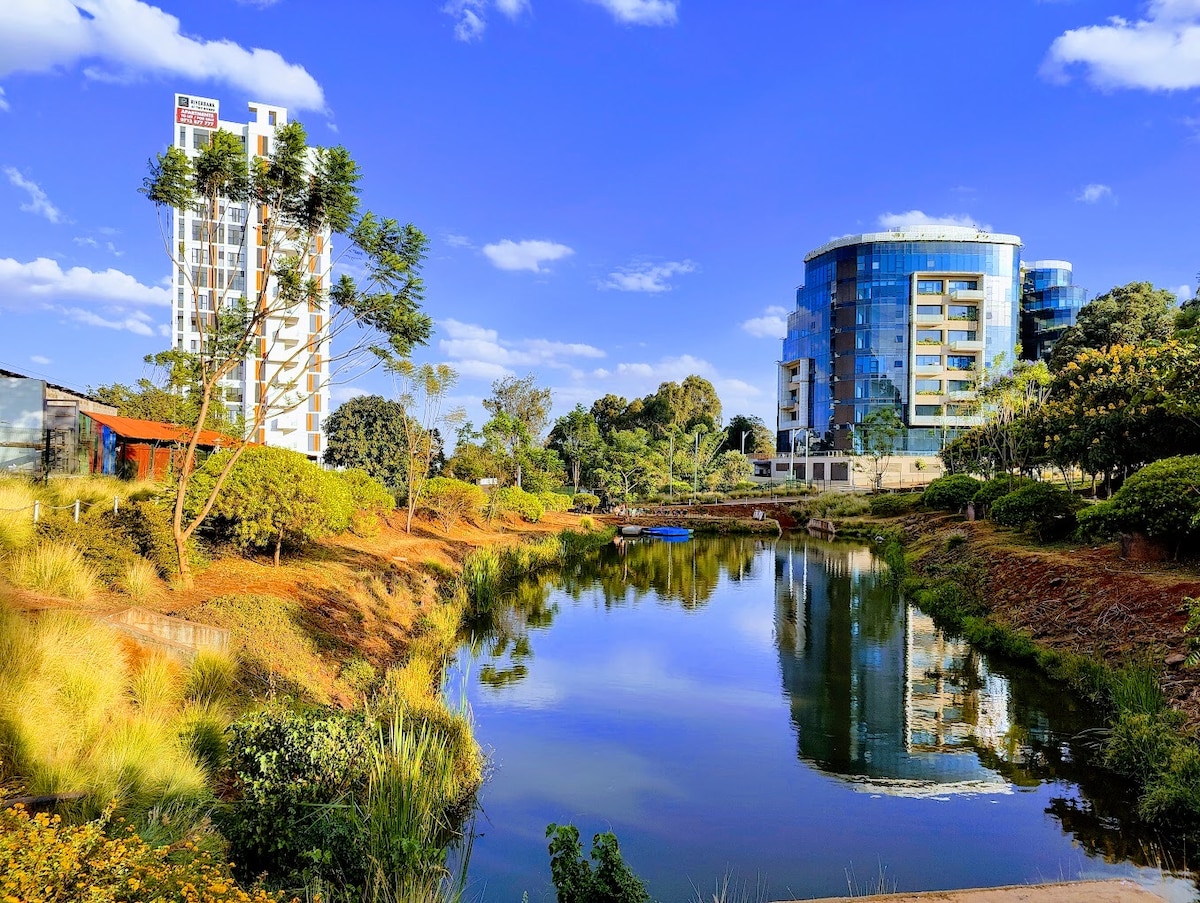 The image captures a serene body of water surrounded by lush greenery and modern buildings. Clear blue skies reflect on the calm surface of the water, with trees framing the scene and contemporary structures standing in the background, enhancing the tranquil environment.