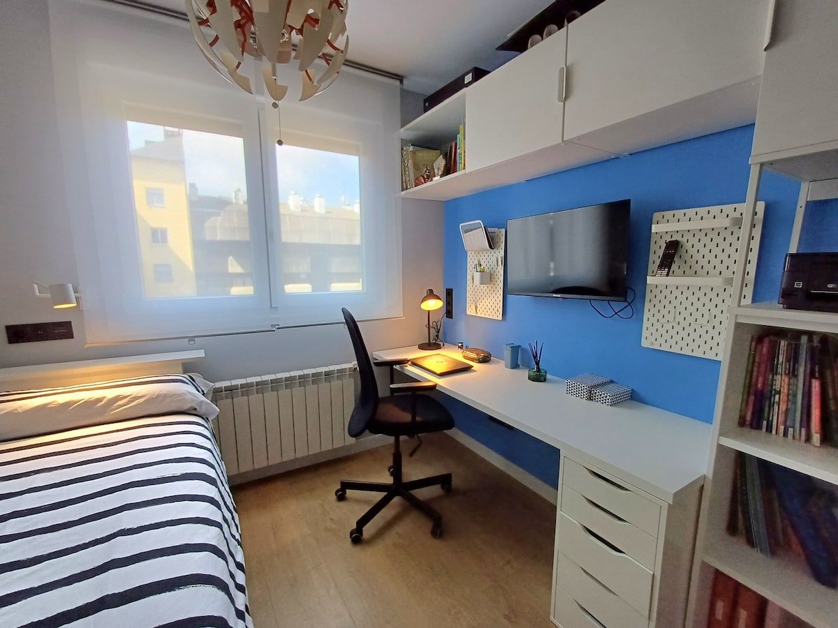 A well-organized bedroom features a spacious bed with striped bedding, a desk with an adjustable chair, and a mounted television. Natural light enters through two windows, enhancing the blue wall behind the desk. Storage shelves and accessible books create a functional living space.