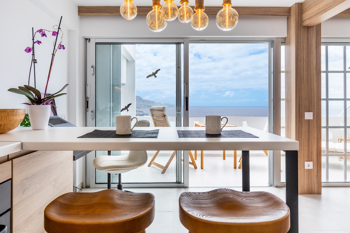 A modern kitchen area is highlighted by a sleek dining table with two chairs and an overhead light fixture featuring multiple bulbs. Large sliding glass doors provide a view of the ocean and outdoor seating, creating a light-filled atmosphere.
