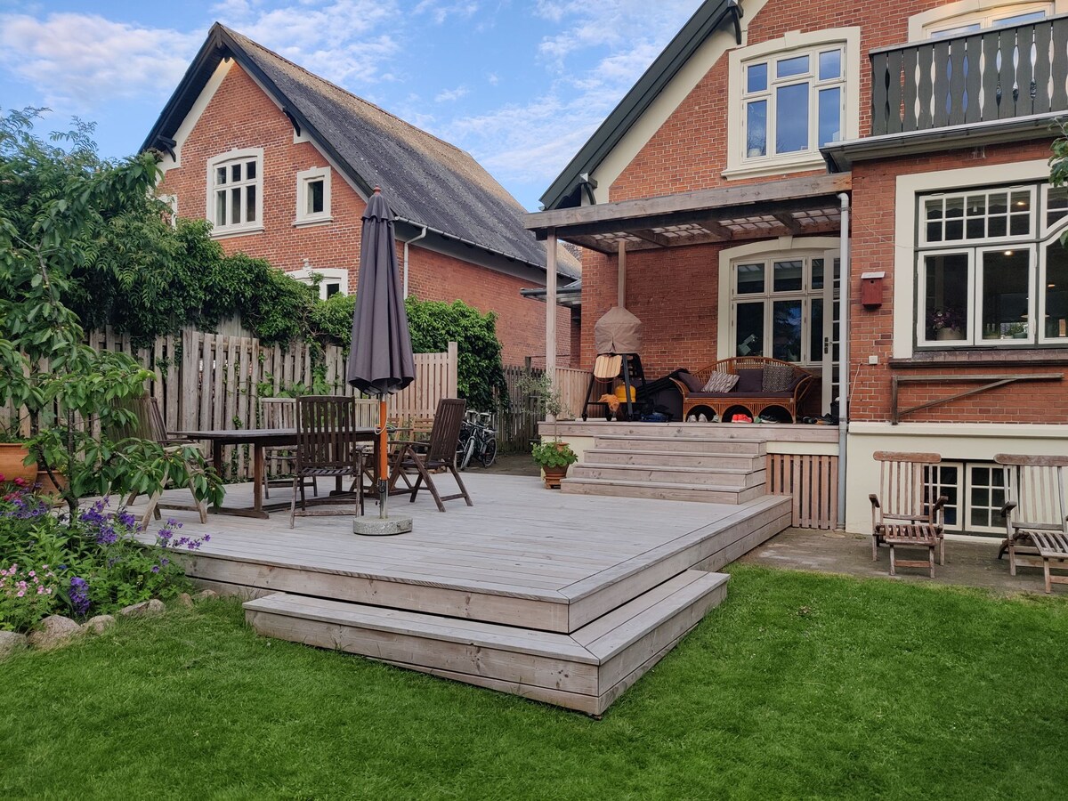 A spacious wooden terrace is seen at the back of the house, featuring a dining table with umbrella and multiple seating areas. Lush green grass surrounds the terrace, complemented by flowering plants. The exterior of the house is brick, showcasing large windows and a balcony.
