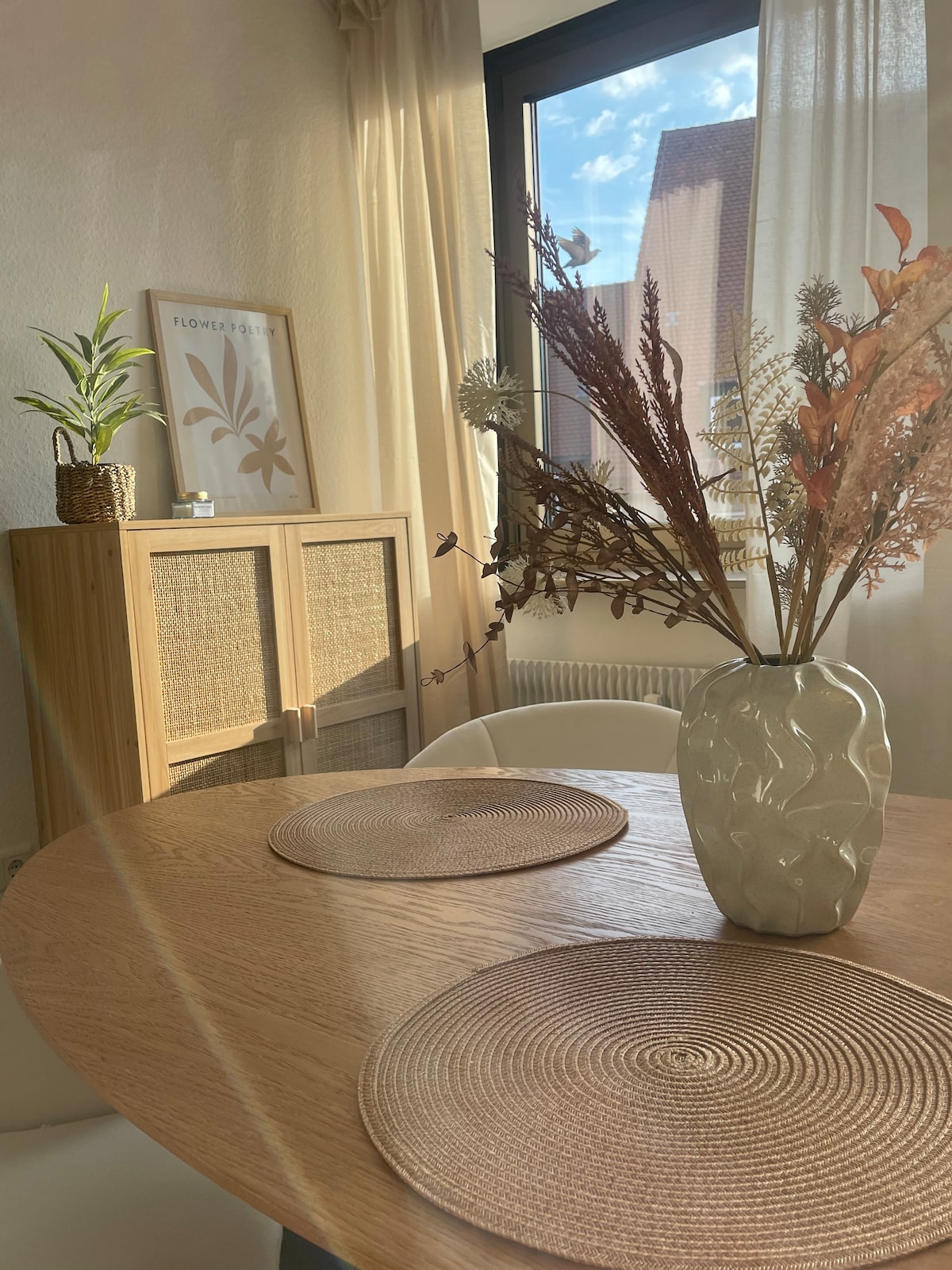 A cozy dining area features a round wooden table with woven placemats, accented by a decorative vase filled with dried flowers. Natural light streams in through a window, highlighting the soft textures of the space. In the background, a cabinet adds an elegant touch.