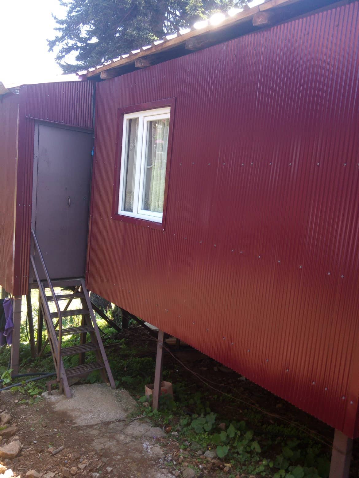 A small structure features a red corrugated metal exterior with a single white window. Stairs lead up to a metal door, providing access to the elevated space. Natural surroundings frame the building, offering a sense of seclusion.