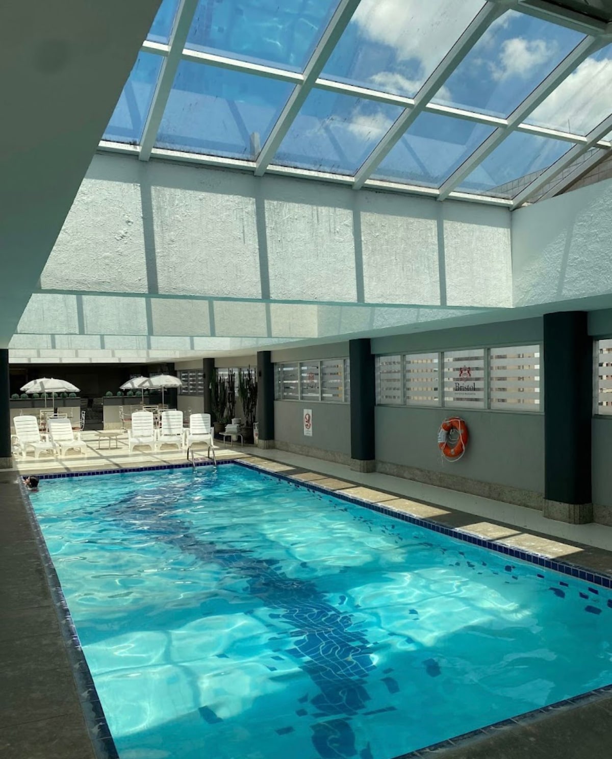 An indoor pool area is visible, featuring clear blue water reflecting natural light from a skylight above. Sun loungers are positioned along the side, and safety equipment is mounted on the wall. The space is framed by large windows, providing an open feel.