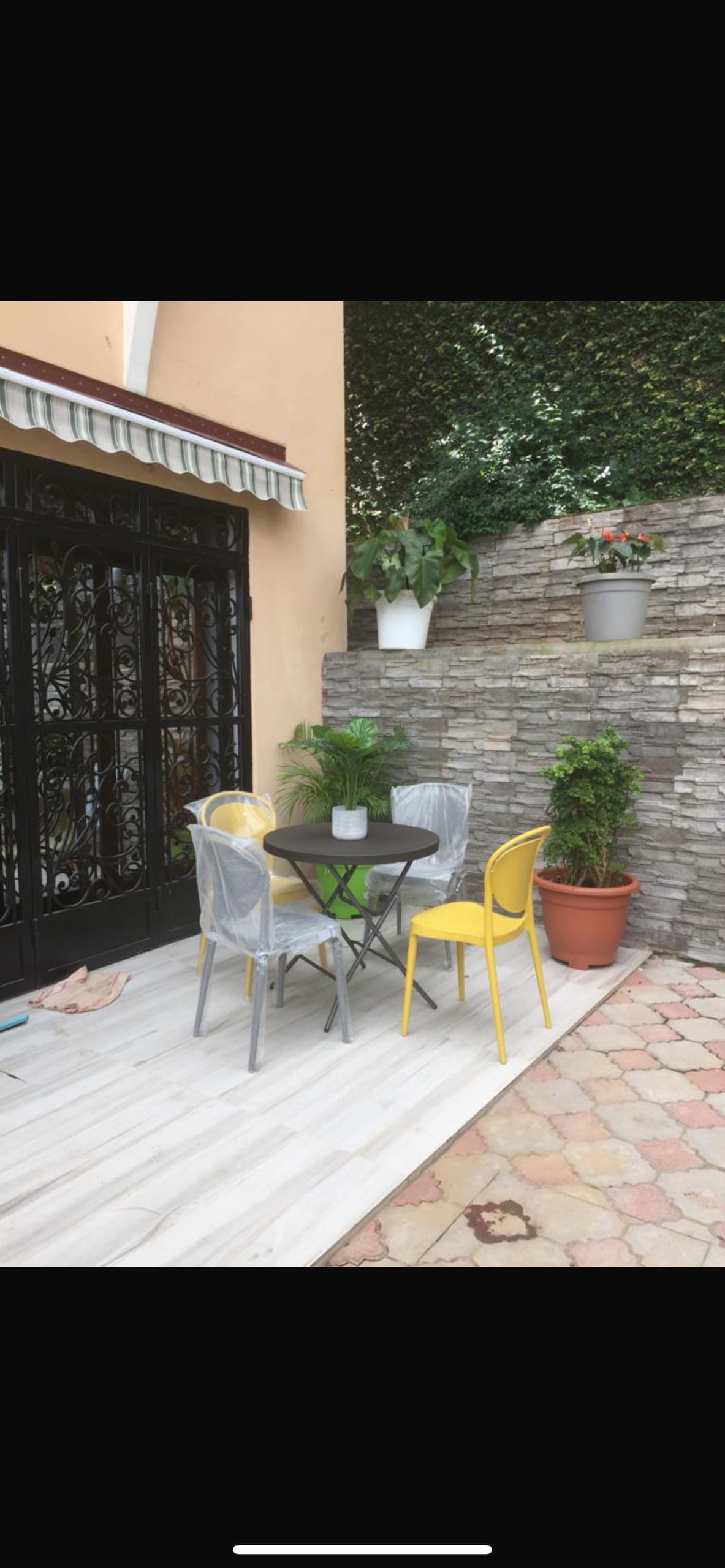 An outdoor seating area is presented with a small round table surrounded by two yellow chairs and two transparent chairs. Potted plants are situated nearby, and a stone wall adds texture to the space, providing a serene environment for relaxation.