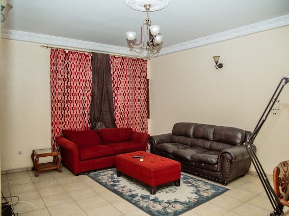 The living area features a red upholstered sofa and a brown leather couch, arranged around a rectangular coffee table. A rug with floral patterns is placed on tiled flooring, under a chandelier that provides ambient lighting. Decorative curtains frame the windows.