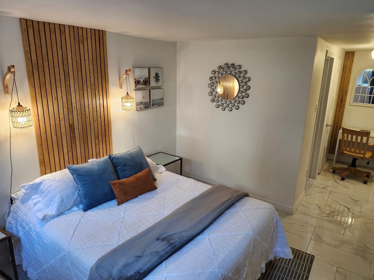 A spacious bed is flanked by two woven pendant lights, providing soft illumination. A decorative wall features wooden paneling, while a mirror and framed art add visual interest. A desk for workspace is situated nearby, contributing to the functional layout of the room.