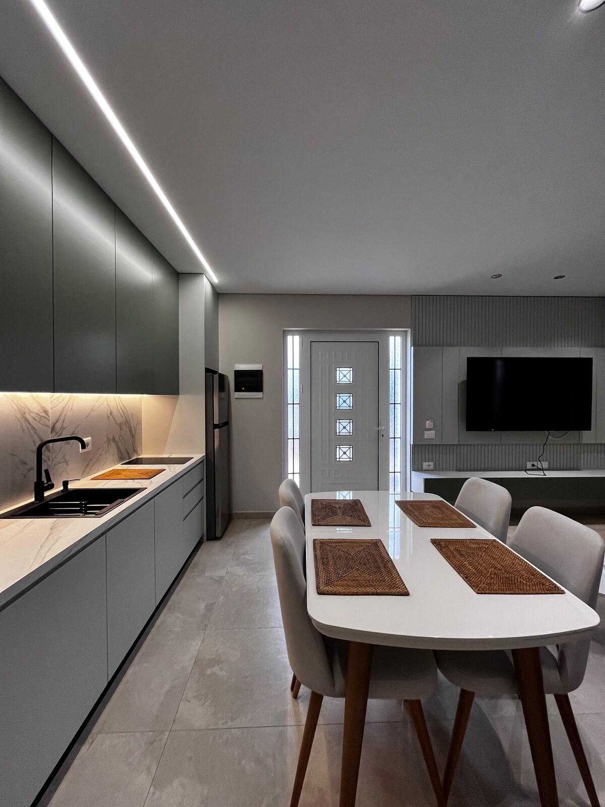 A modern kitchen features sleek grey cabinetry and a white countertop. A dining table with light grey chairs is set with placemats, creating a space for shared meals. Natural light filters in from a door leading to the outdoors, complementing the neutral tones.