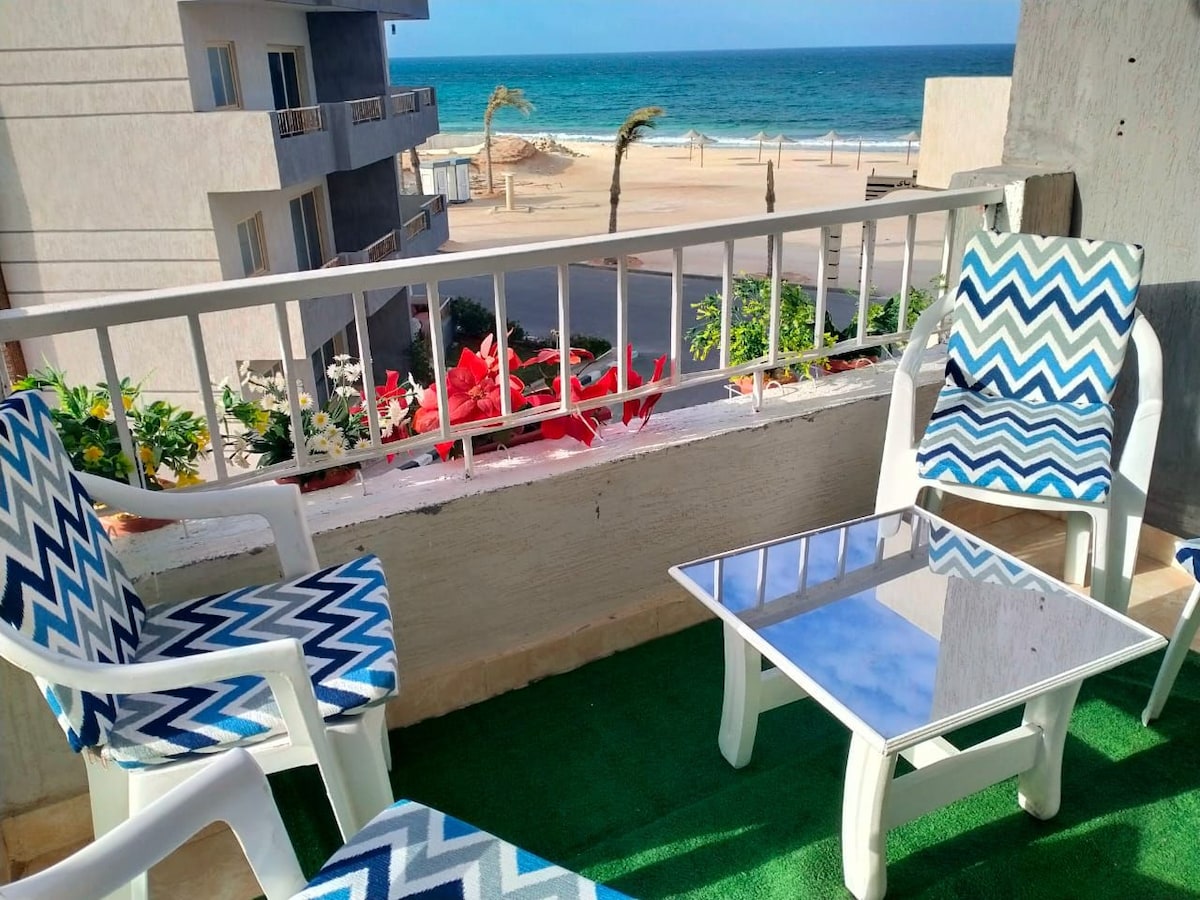 A small balcony is furnished with two patterned chairs and a glass-top table. A view of the ocean is visible beyond the railing, complemented by potted plants and a sandy beach in the distance. The space is bright and open, inviting relaxation.