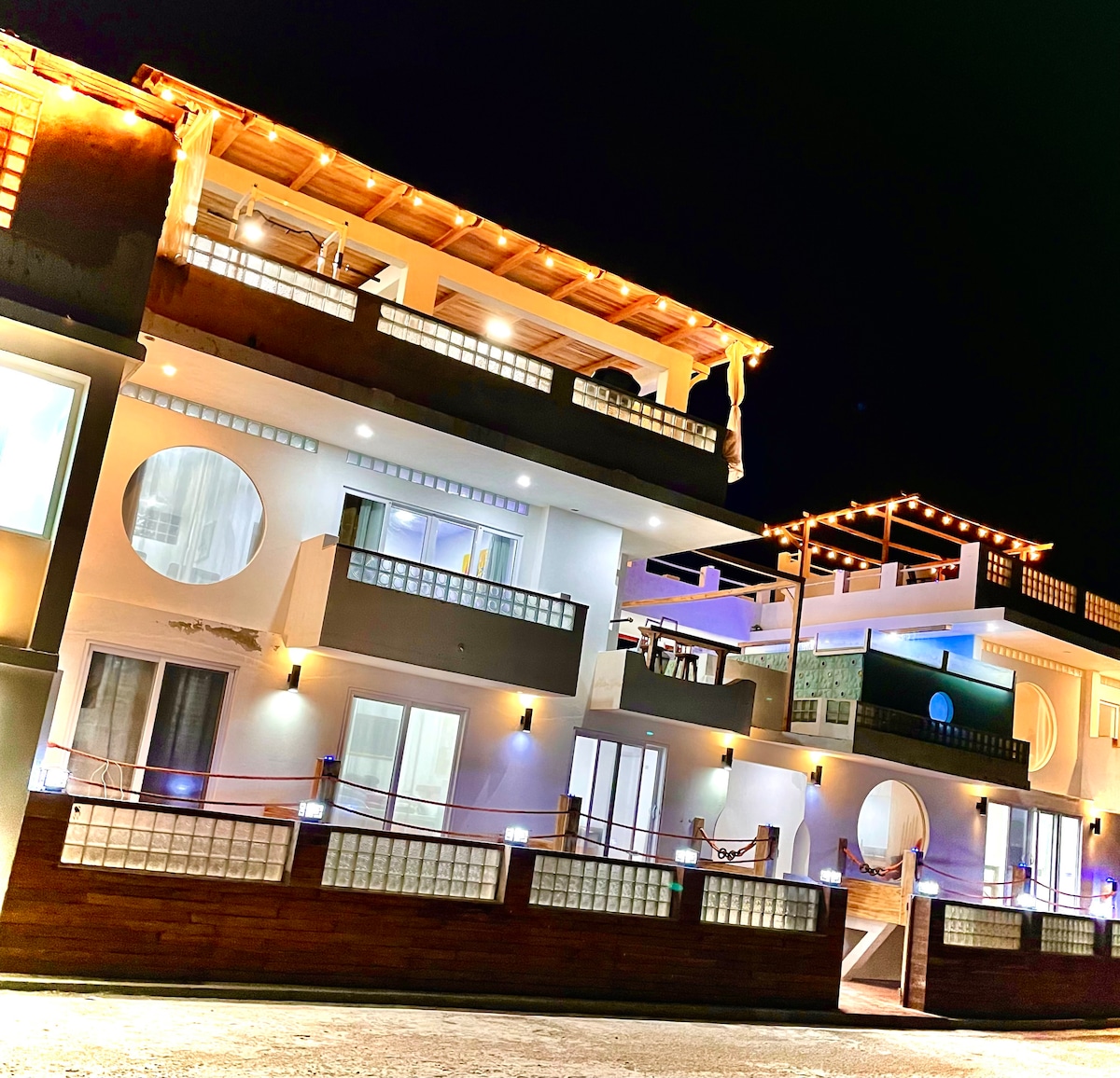 The multi-story Casa Barco is illuminated at night, showcasing its unique architectural features and smooth lines. Each level includes balconies and large windows, allowing for a blend of indoor and outdoor living. The inviting atmosphere is enhanced by warm lighting along the terrace.