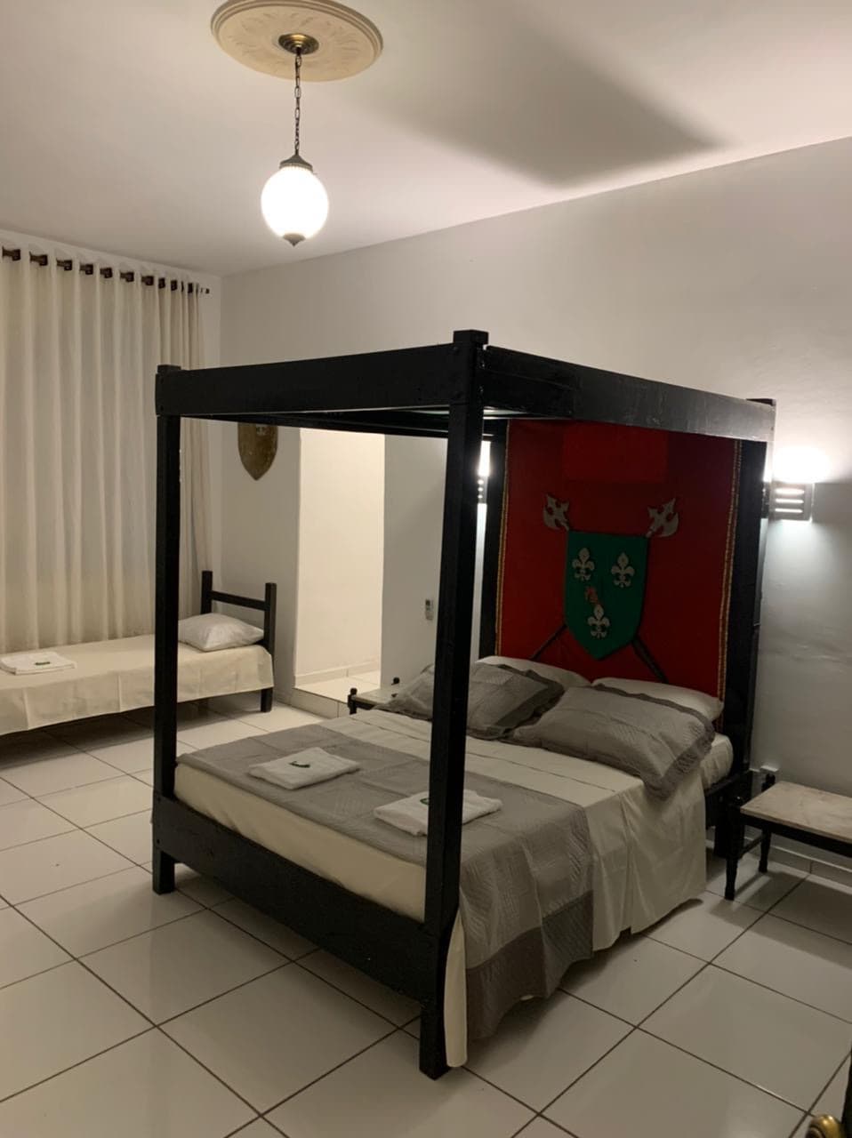 The room features a four-poster bed with a decorative medieval backdrop. A twin bed is positioned nearby, contributing to the spacious layout. Light-colored linens and towels are neatly arranged, while large windows dressed with sheer curtains provide natural light to the space.