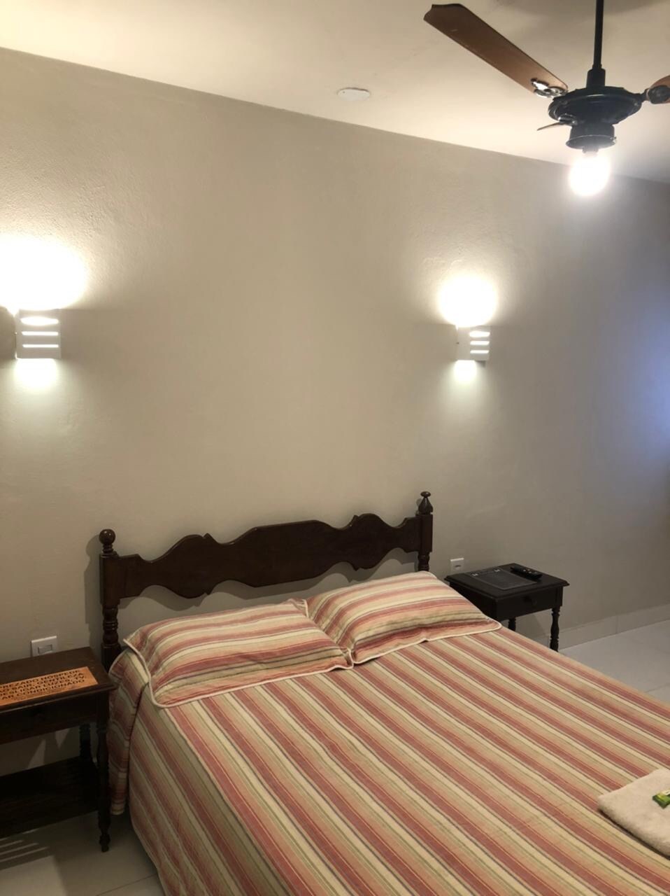 The bedroom features a spacious bed with a striped duvet, flanked by two wall-mounted lamps. A small bedside table is positioned next to the bed. Light from a nearby window softly illuminates the neutral-colored walls.