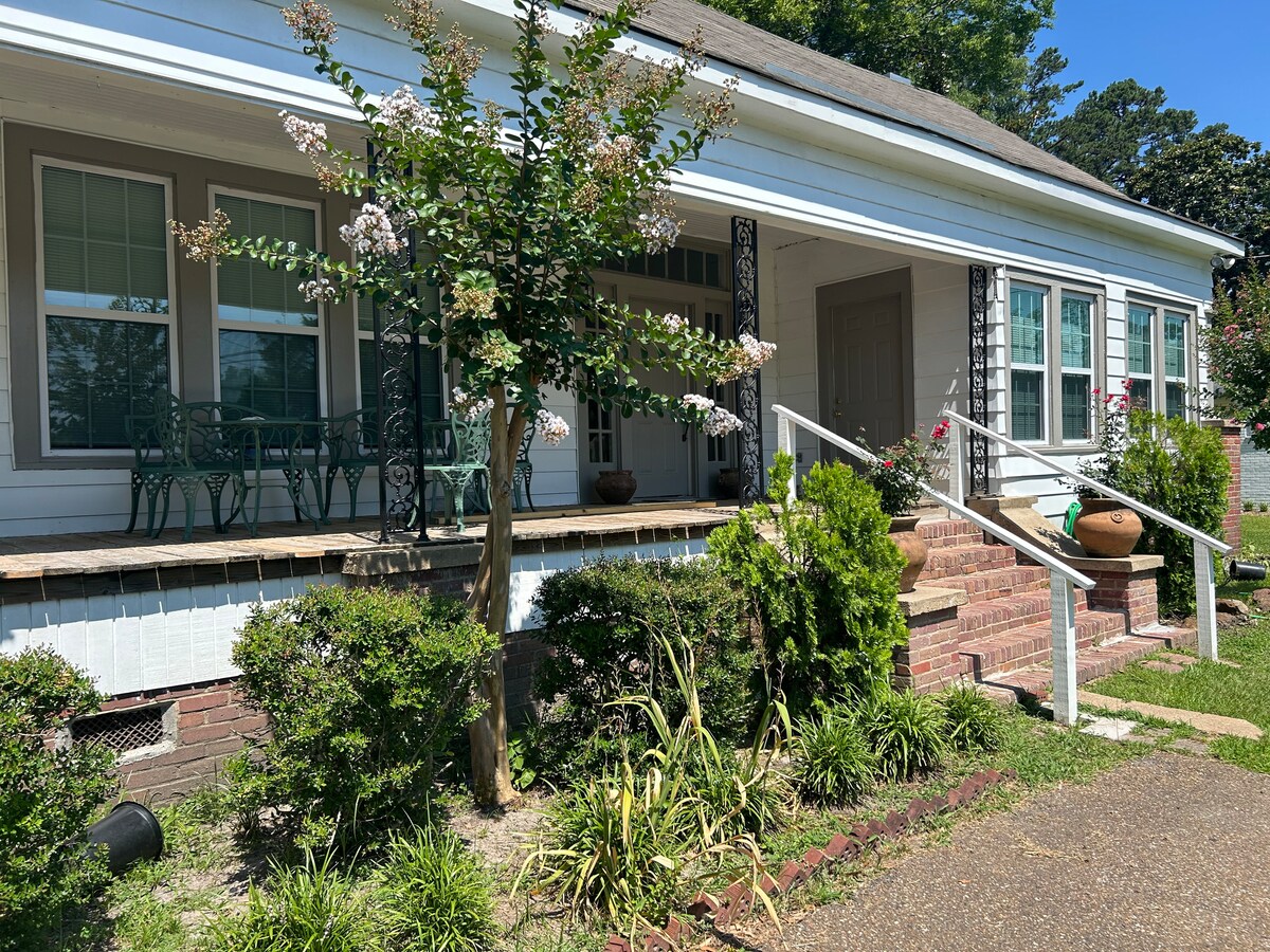 The exterior of the house features a charming porch with decorative iron railings and a seating area. Lush greenery surrounds the home, including shrubs and flowers, complementing the classic architecture. The entrance is framed by large windows that invite ample natural light.
