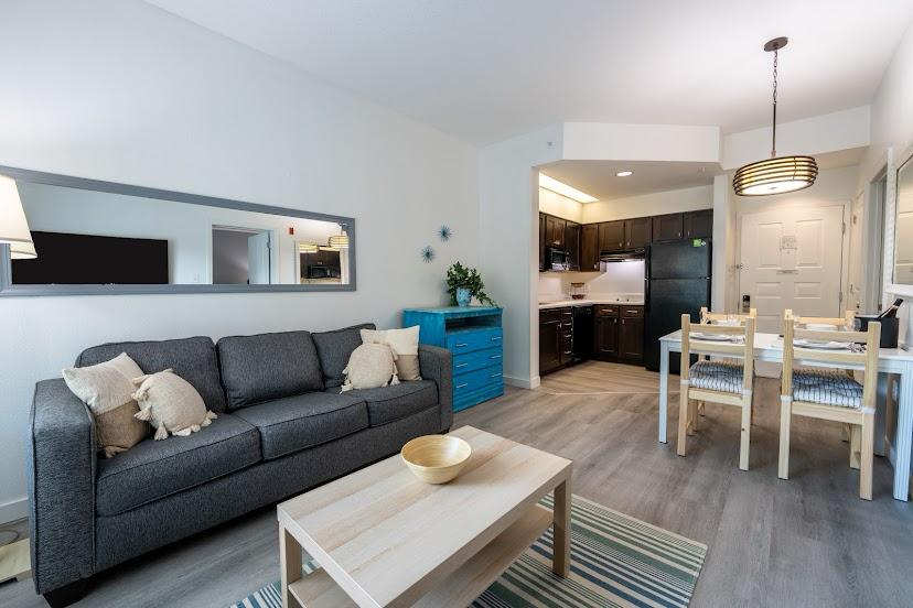 The living area features a gray sofa, enhanced by decorative pillows and accentuated by a mirrored wall. A wooden coffee table is centered on a striped area rug, while a dining table with four chairs occupies the space, with a blue dresser visible in the background.
