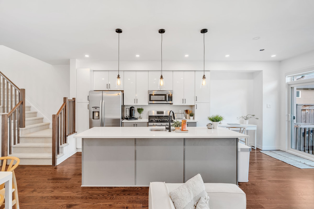 A modern kitchen features a large central island with seating. Stainless steel appliances, including a refrigerator and microwave, are visible. Pendant lights hang above, illuminating the sleek design. A staircase leads to the upper level, while a door offers access to the outdoor space.