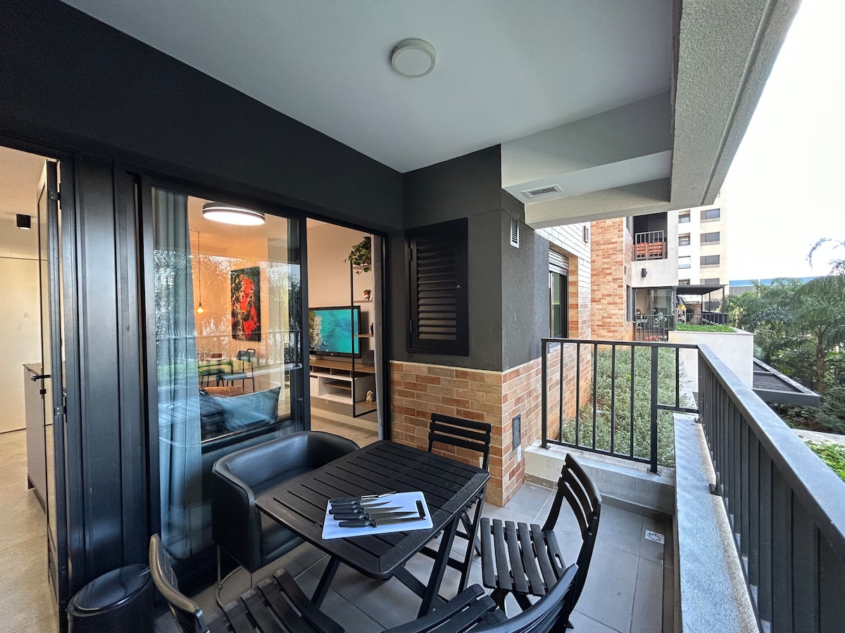 A balcony area is shown, featuring a dining table with four chairs. A view of greenery is visible from the space, along with an outdoor seating arrangement. Large glass doors separate the balcony from the living area, allowing natural light to flow inside.