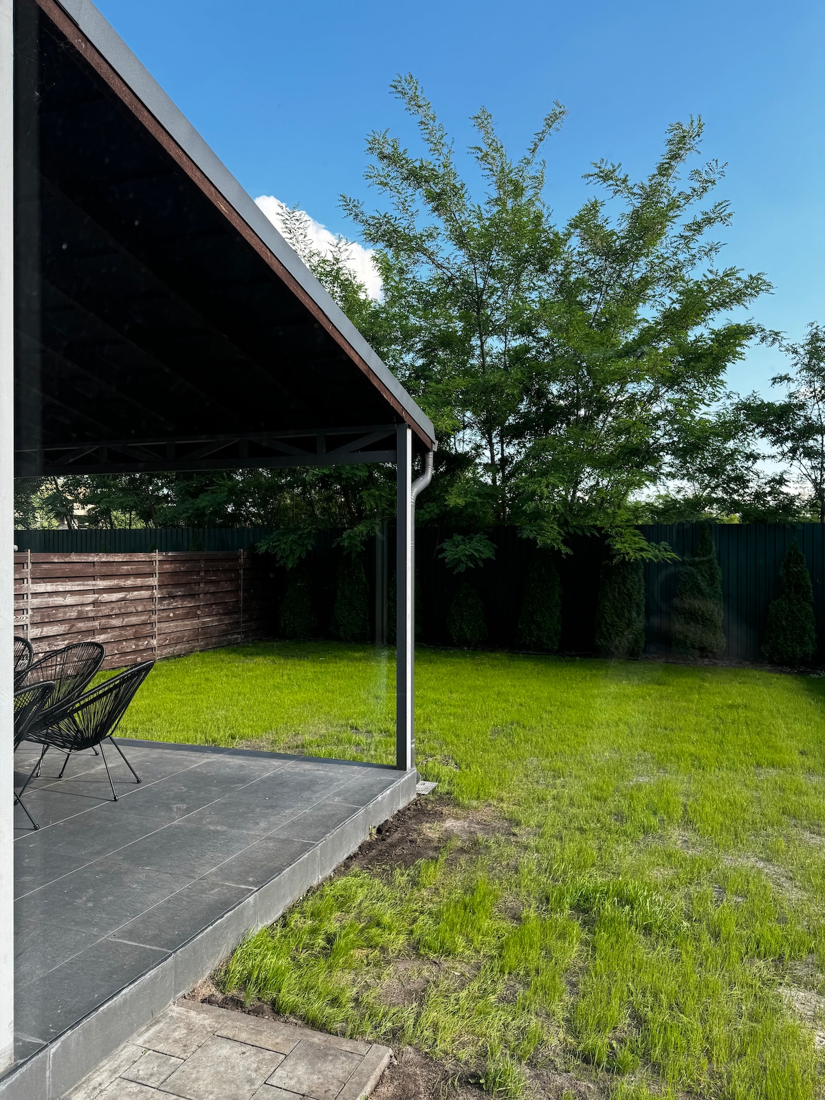 A covered patio area is visible, featuring a set of black chairs around a table. The neatly maintained green lawn extends beyond, bordered by trees and shrubs, creating a serene outdoor space. The clear blue sky adds to the inviting atmosphere.