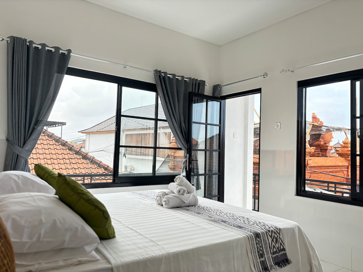 A large room features a spacious bed with white linens and green decorative pillows. Large windows with black frames open to a view of the outside. Light-colored curtains are neatly arranged, allowing for natural light to fill the space while highlighting the room's clean lines.