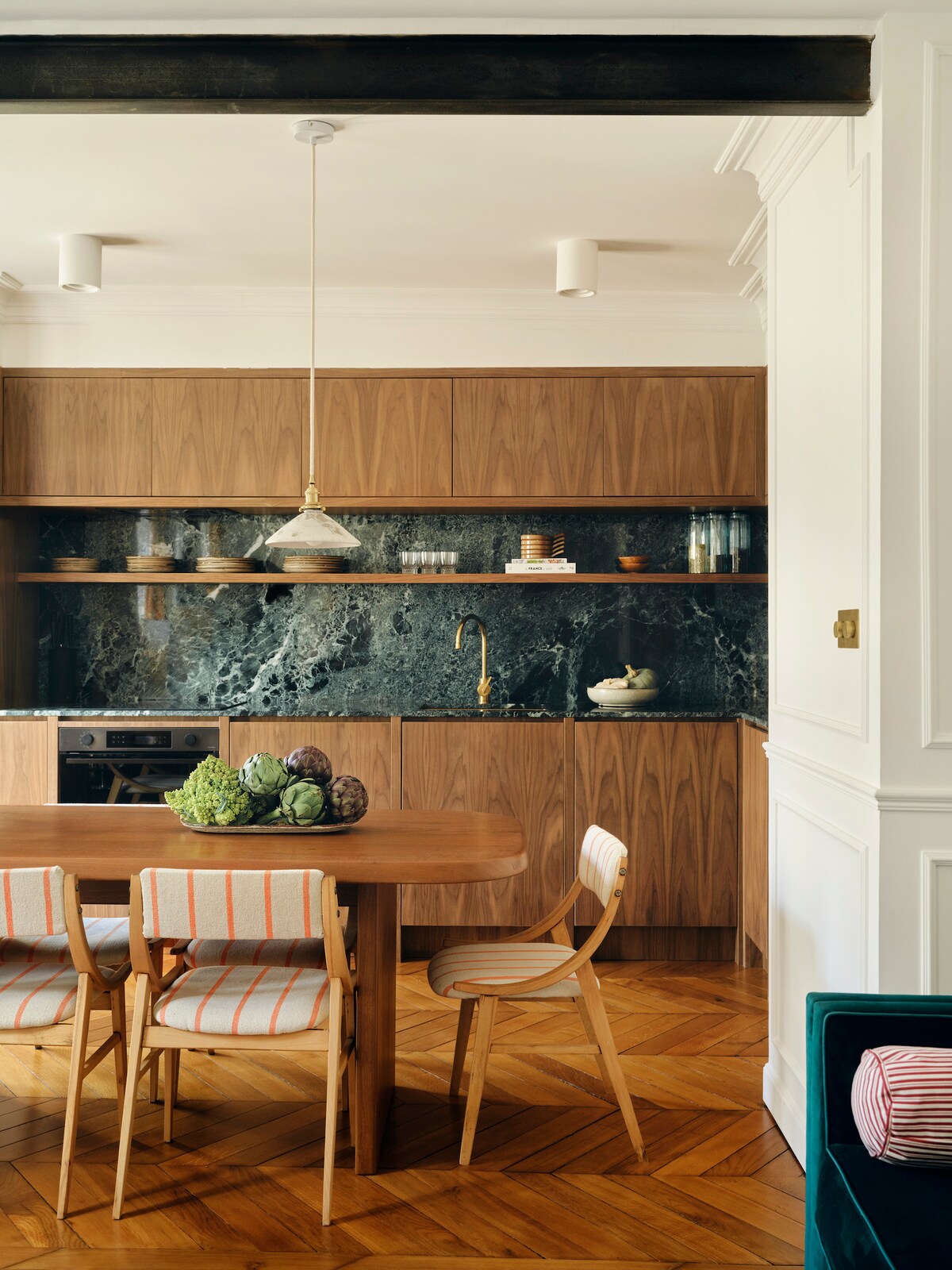 A modern kitchen features rich wooden cabinetry and a dark marble backsplash. A round dining table is surrounded by four chairs, including two with striped upholstery. Fresh vegetables are artistically arranged on the table. The space is illuminated by a hanging pendant light.