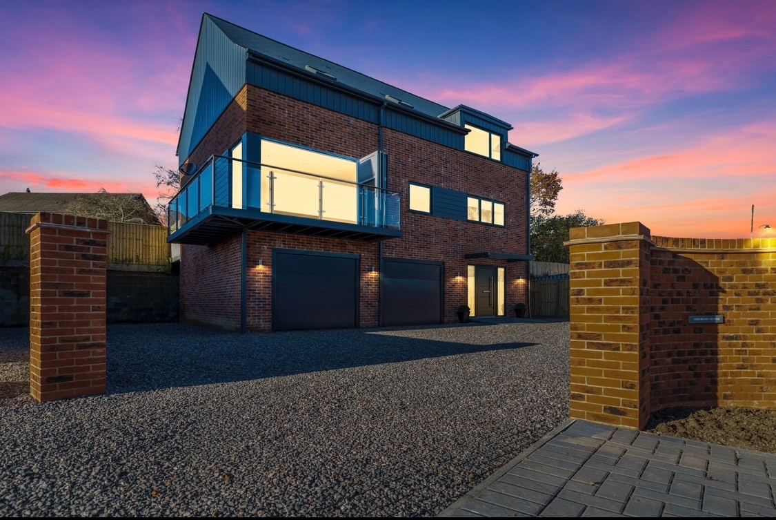 A modern, three-story brick house stands illuminated at twilight, featuring large glass windows and a front balcony. The property is surrounded by gravel parking and presents an appealing façade with two garage doors. Warm evening hues paint the sky, enhancing the setting.