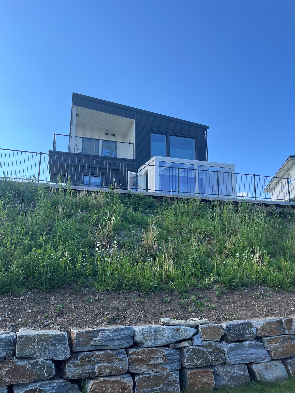 A modern house is set on a hillside, featuring a large glass terrace on the upper level. The exterior showcases a combination of dark wood and white walls, surrounded by greenery. A stone wall borders the property, adding a natural contrast to the landscape.