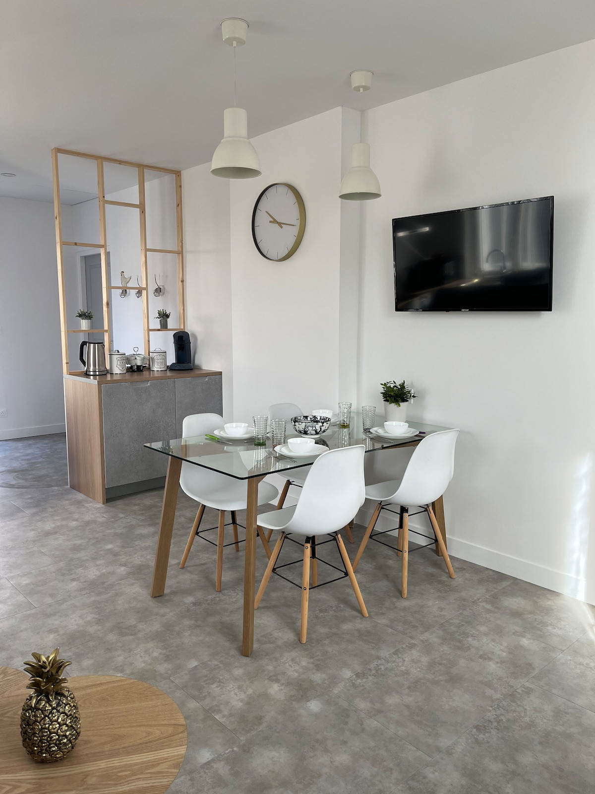 An open dining area features a glass-top table surrounded by four white chairs. A wall-mounted television is present, along with a clock above. The kitchen is visible in the background, showcasing a sleek and modern design with essential appliances.