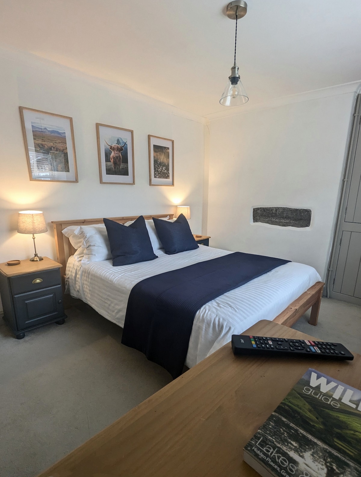 A cozy bedroom features a spacious bed adorned with navy-blue pillows and a light duvet. Two bedside lamps provide soft lighting, complementing framed images on the wall. A wooden desk holds a remote control and a guidebook. A muted palette enhances the tranquil atmosphere.