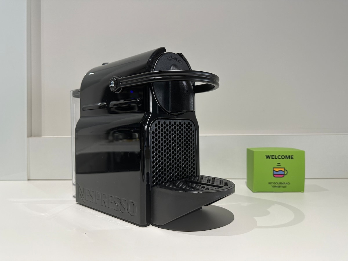 A Nespresso coffee machine is displayed on a white countertop, featuring a sleek black design with a drip tray. A green welcome kit is positioned nearby, highlighting starter items for guests.