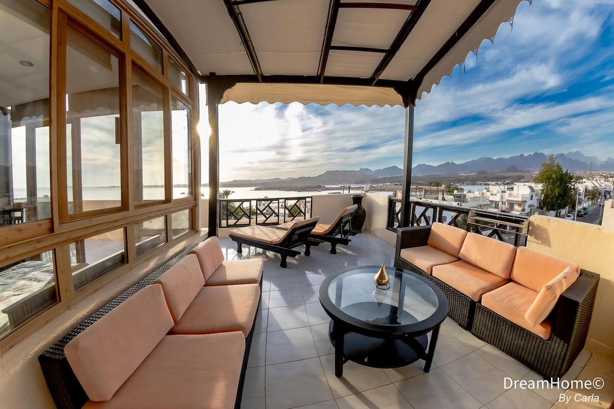 A cozy outdoor seating area features a circular glass table surrounded by comfortable couches. The view of the sea and mountains is visible through large windows, creating an inviting space for relaxation. Natural light enhances the tranquil atmosphere of the area.
