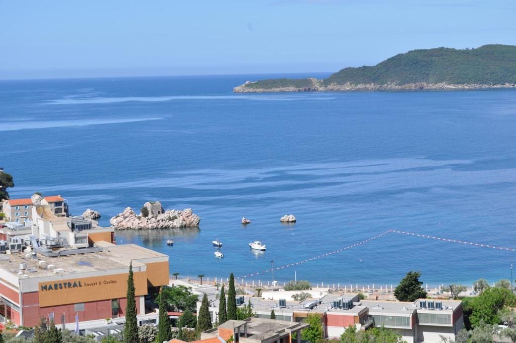 A panoramic view of a calm sea is captured, displaying clear blue waters and several boats gently floating. The coastline features a mix of rocky formations and sandy areas, with trees and buildings lining the foreground. The distant hills provide a serene backdrop.