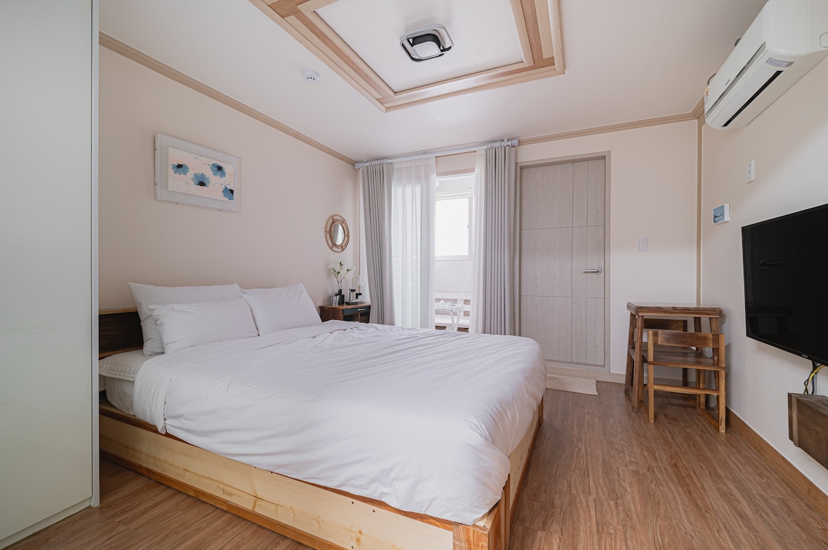 A spacious bedroom is designed with light-colored walls and wooden accents. A large bed is centered in the room, with crisp white linens. Natural light streams through a partially draped window, illuminating the space. A minimalist wooden table is positioned beside the bed.