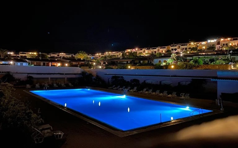 A spacious outdoor pool is illuminated at night, showcasing vibrant blue water. Surrounding the pool, lounge chairs are arranged neatly along the patio. Dim lighting accentuates the landscape, with softly lit buildings rising on the hillside, creating a peaceful ambiance.