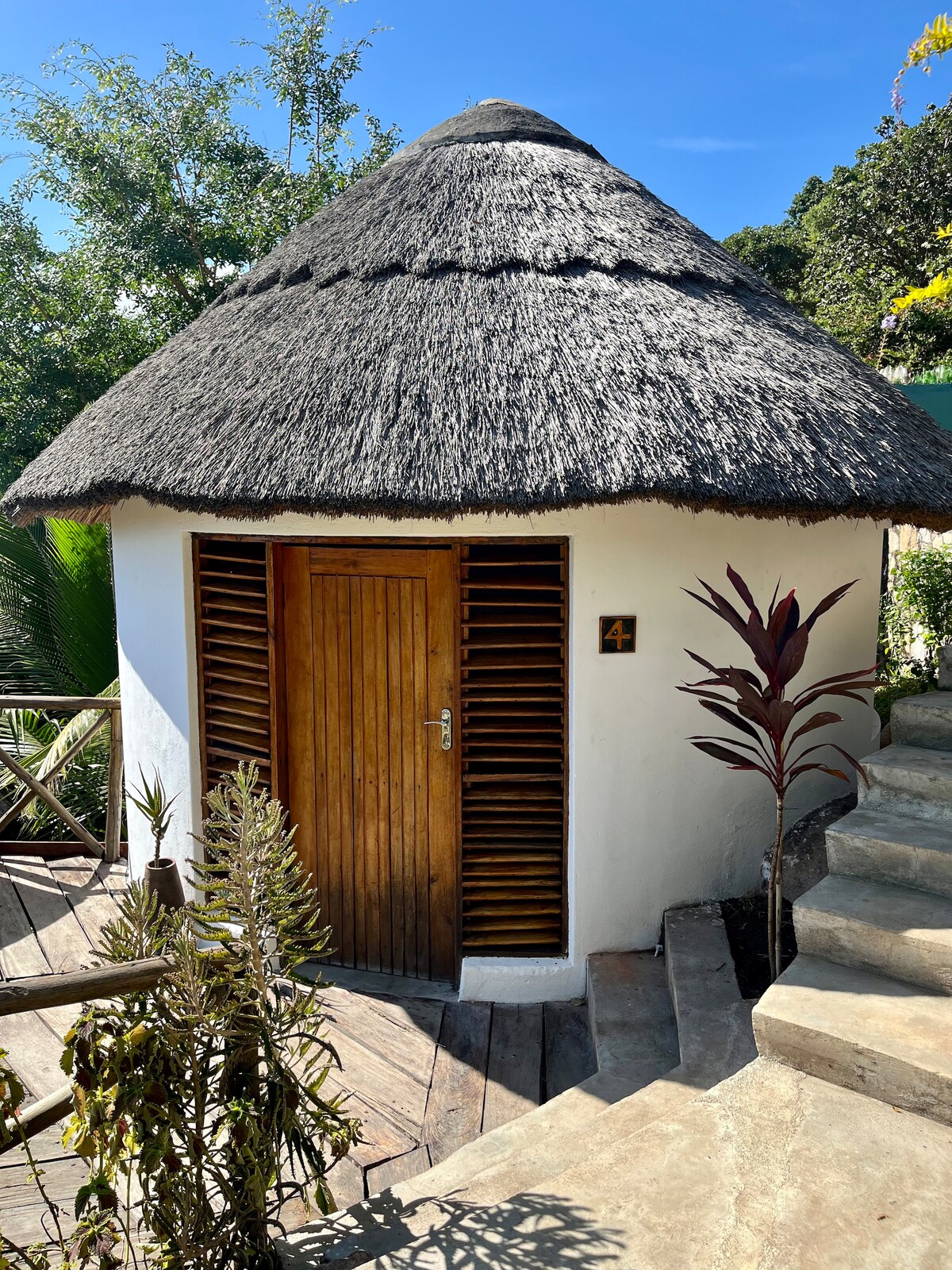 A round, thatched-roof structure is shown, with a wooden door featuring horizontal slats. The entrance is framed by lush greenery, and a solid stone pathway leads to the door. A number '4' is displayed next to the door, indicating the room.