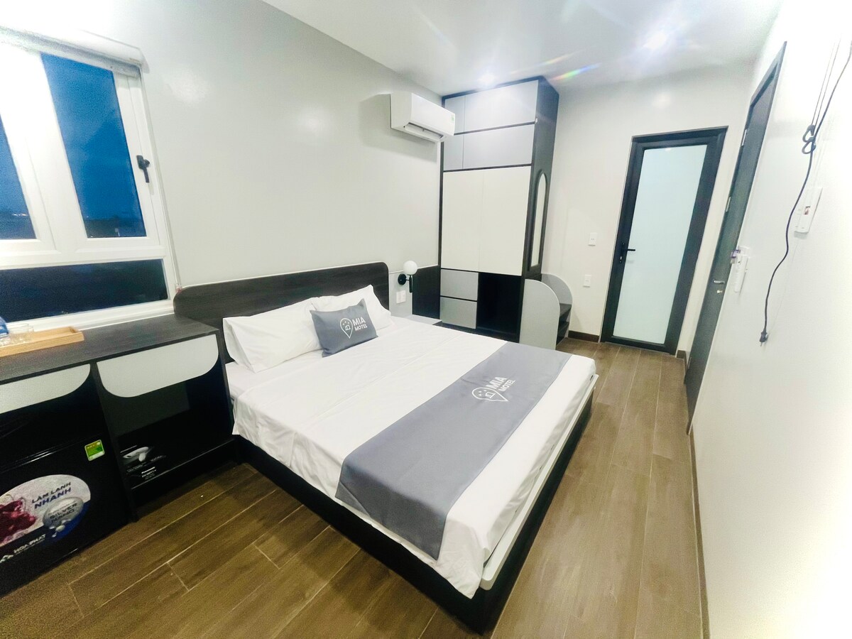 A bedroom features a spacious bed with a light grey bedspread, complemented by a matching headboard. A sleek black and white wardrobe is present, along with a small bedside table. Natural light enters through a window, and a modern air conditioning unit is installed on the wall.