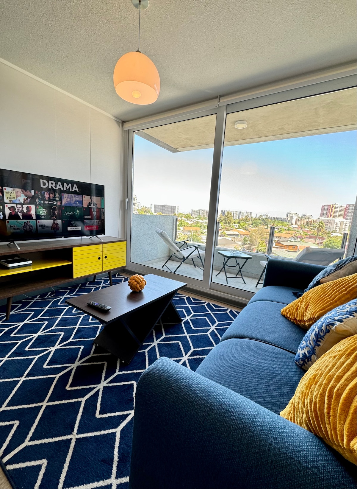 A cozy living area features a blue sofa adorned with yellow pillows, positioned near a large glass window that offers views of the cityscape. A modern TV stand with a yellow finish supports a flat-screen TV. A geometric rug in shades of blue and white adds a vibrant touch.
