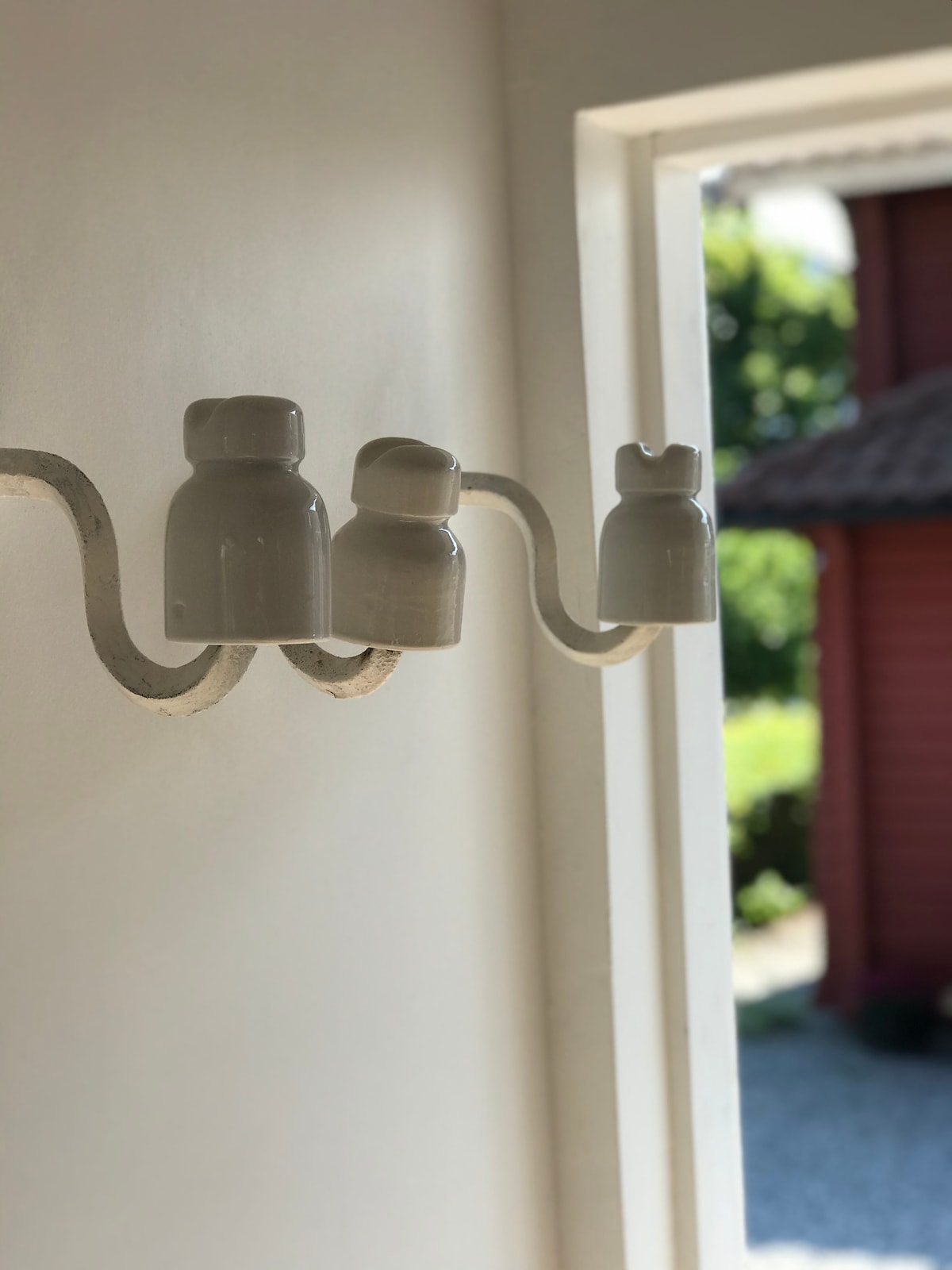 A wall sconce featuring three white ceramic fixtures is visible, mounted on a curved metal arm. Soft natural light filters through a nearby doorway, casting gentle shadows. A glimpse of greenery and a red structure is seen in the background.