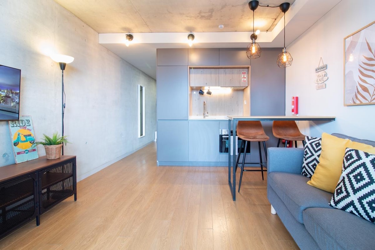 An open living area features a modern kitchen with sleek cabinetry and a breakfast bar with two wooden stools. A comfortable grey sofa is accompanied by a decorative pillow. Soft lighting is provided by pendant fixtures and a floor lamp, creating a bright and inviting space.