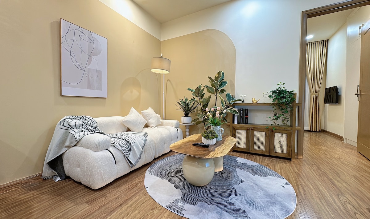 A cozy living area features a light-colored sofa adorned with soft pillows, arranged around a natural wood coffee table. A circular rug adds texture underfoot. Indoor plants in decorative containers enhance the space, while a wall-mounted TV is positioned near a doorway lined with curtains.