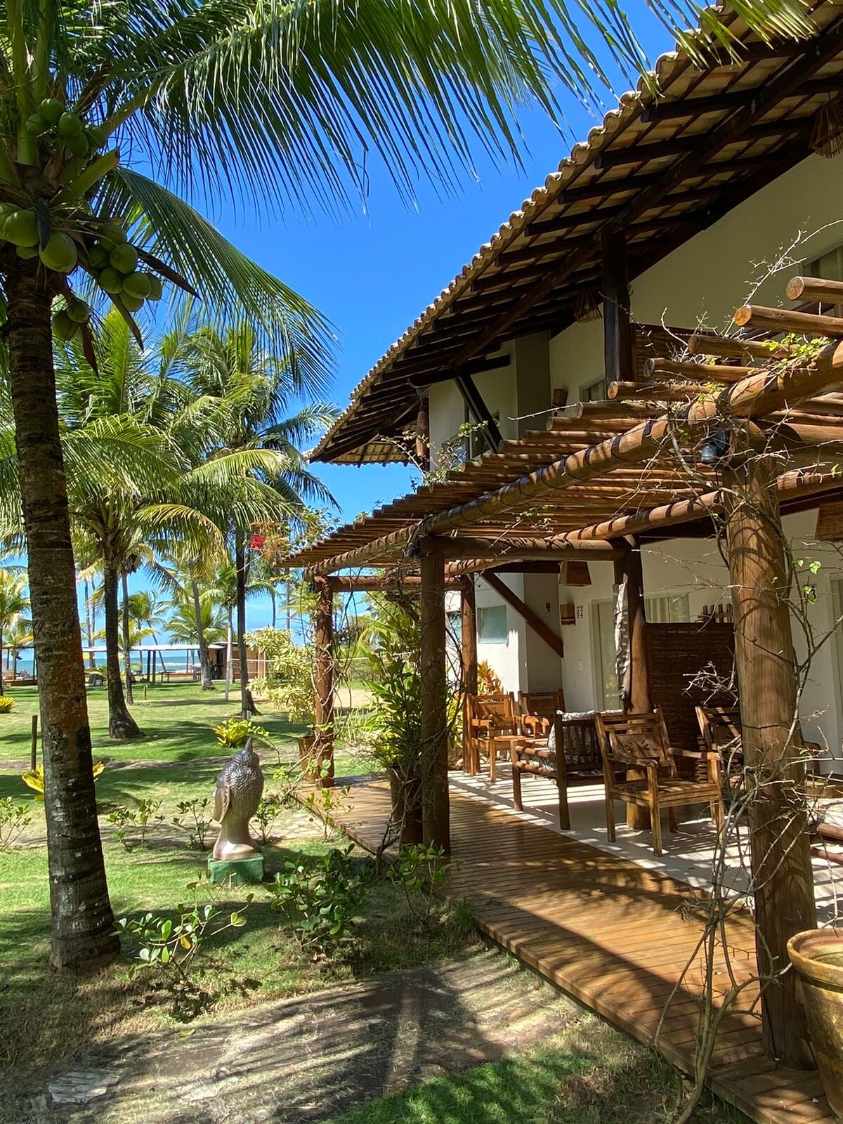 A view of the property's exterior reveals a wooden porch with seating surrounded by tropical palm trees. The structure's natural materials complement the lush garden, while the distant ocean is visible beyond the greenery under a bright blue sky.