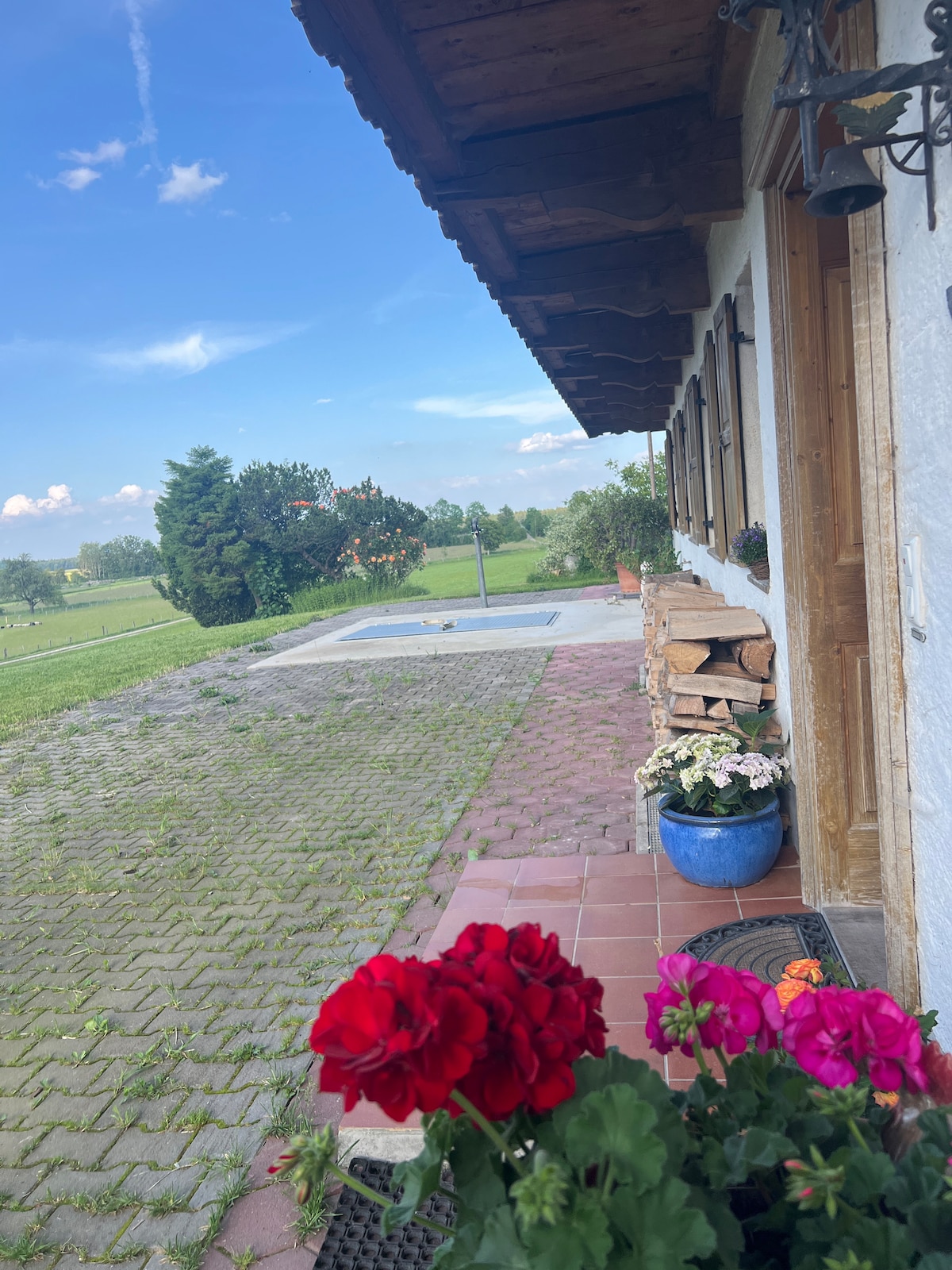 The image captures a charming patio area with vibrant flower pots featuring red and pink blooms. A stone pathway leads to a grassy landscape, framed by greenery. A wooden structure offers shade, while a view of an open area is visible beyond the patio.