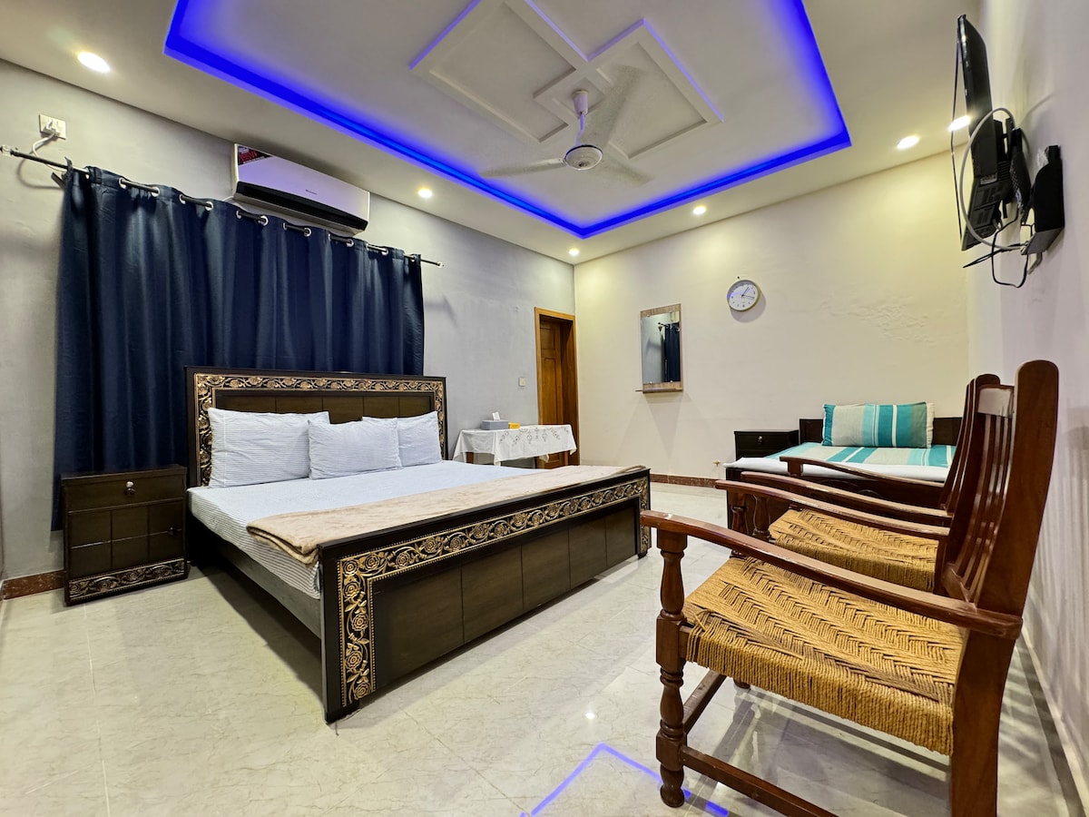 The spacious bedroom features a king-size bed with decorative detailing. A seating area includes two wooden chairs, complementing the modern decor. Soft lighting from the ceiling enhances the ambiance, while dark curtains provide privacy. An air conditioning unit is mounted on the wall for comfort.