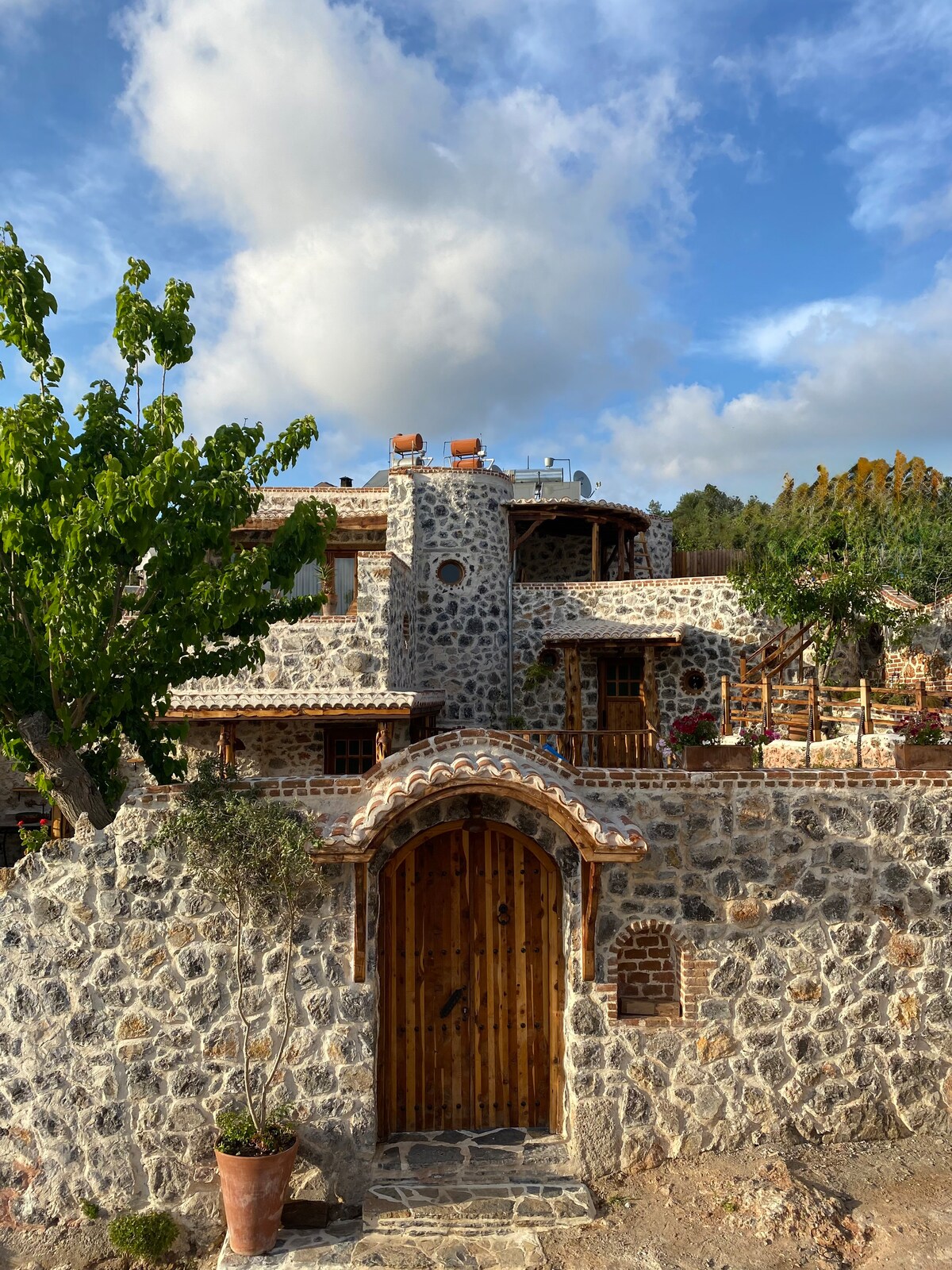 A rustic stone villa is surrounded by lush greenery, with a wooden door that invites guests inside. The architecture highlights traditional elements, and a balcony is visible above the entrance, providing a charming view of the surrounding landscape.