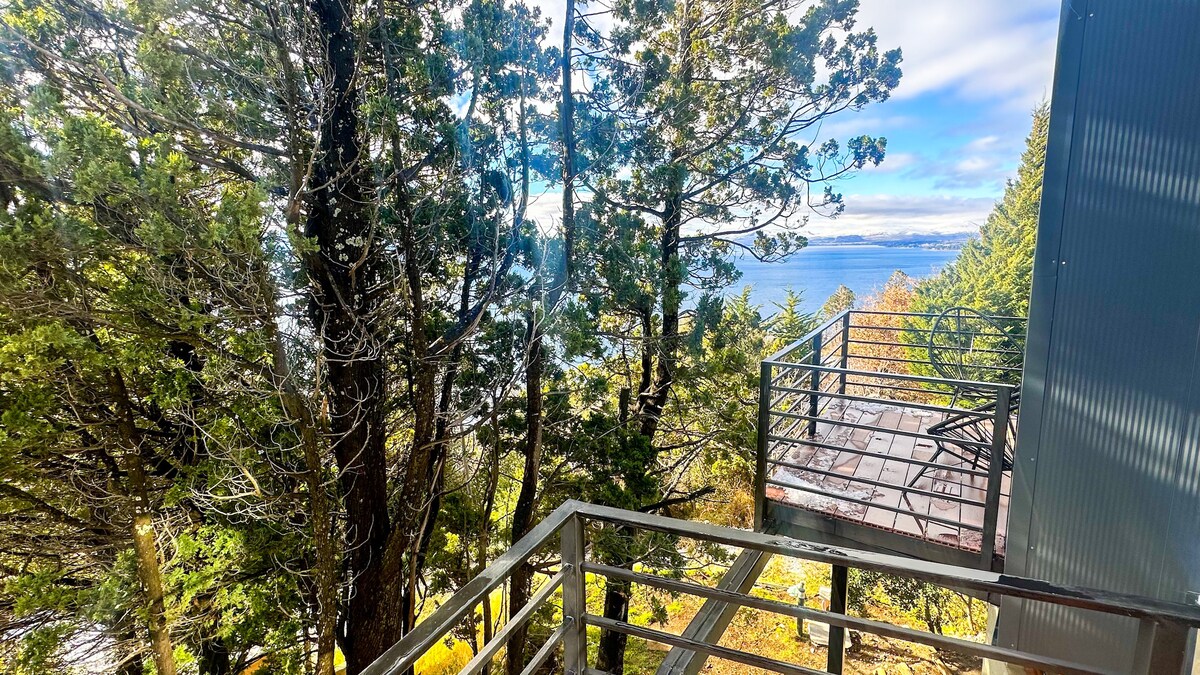 A balcony is visible, surrounded by tall trees offering a view of Lake Nahuel Huapi. A small seating area with a chair and table is situated on the balcony, allowing for relaxation amid nature.
