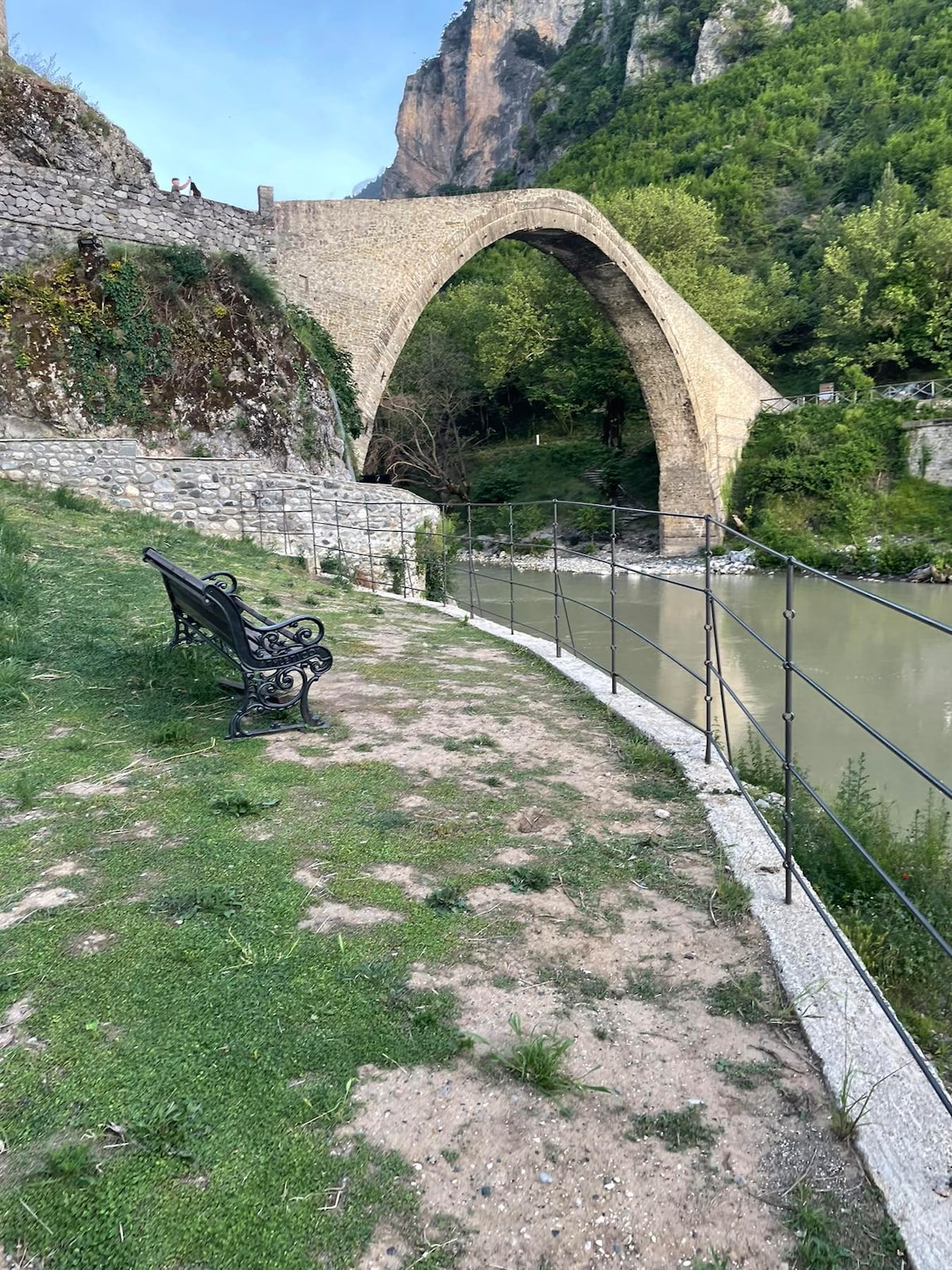 A historic stone arch bridge is prominently featured, spanning a serene river. A black iron bench sits along a grassy bank, providing a space for relaxation and contemplation. Lush greenery surrounds the area, enhancing the natural beauty of the setting.