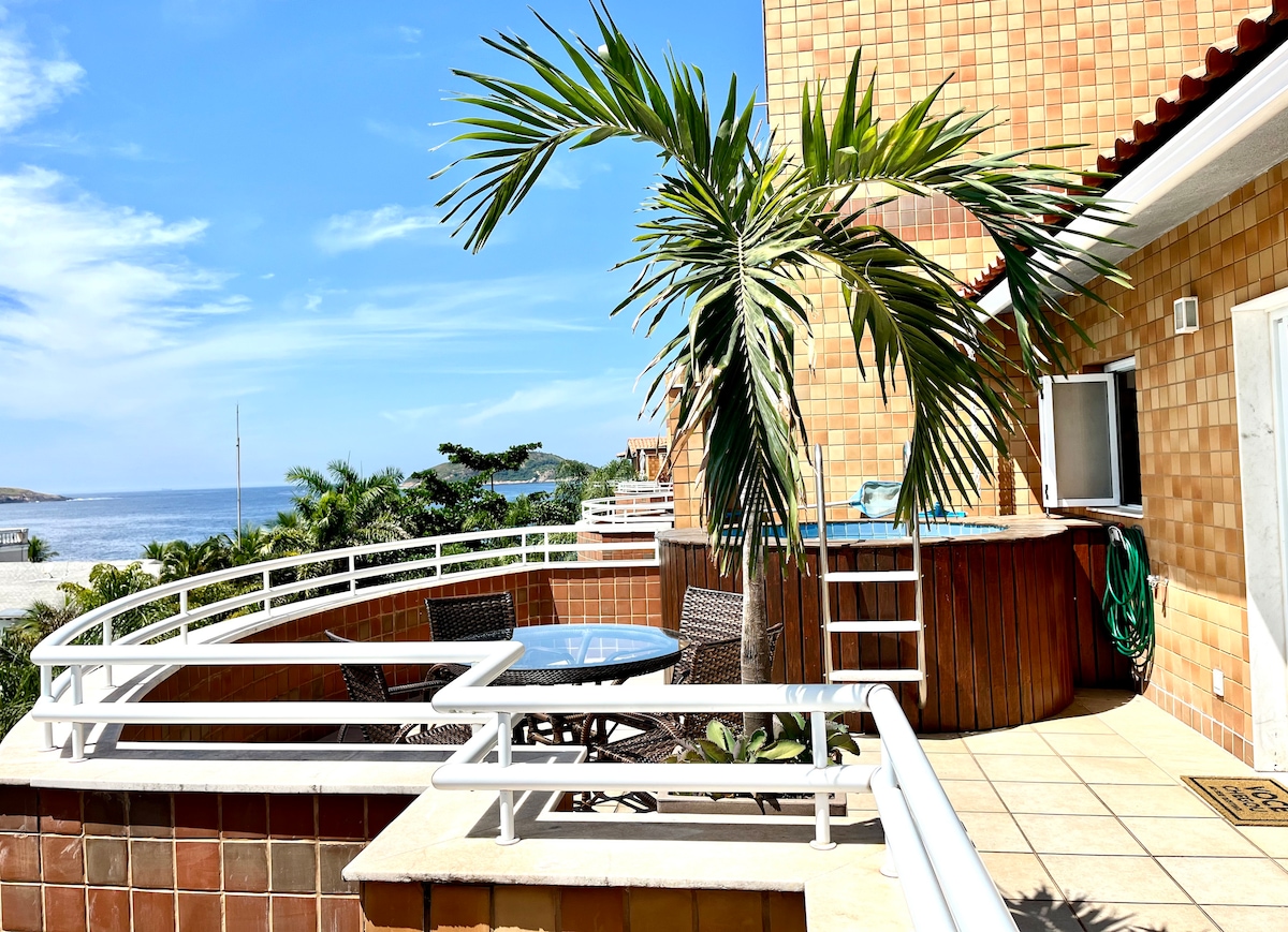 A private outdoor area is featured with a round table and four wicker chairs, surrounded by a low wall. A palm tree provides shade, and a small hot tub is visible. The ocean can be seen in the background under a clear blue sky.
