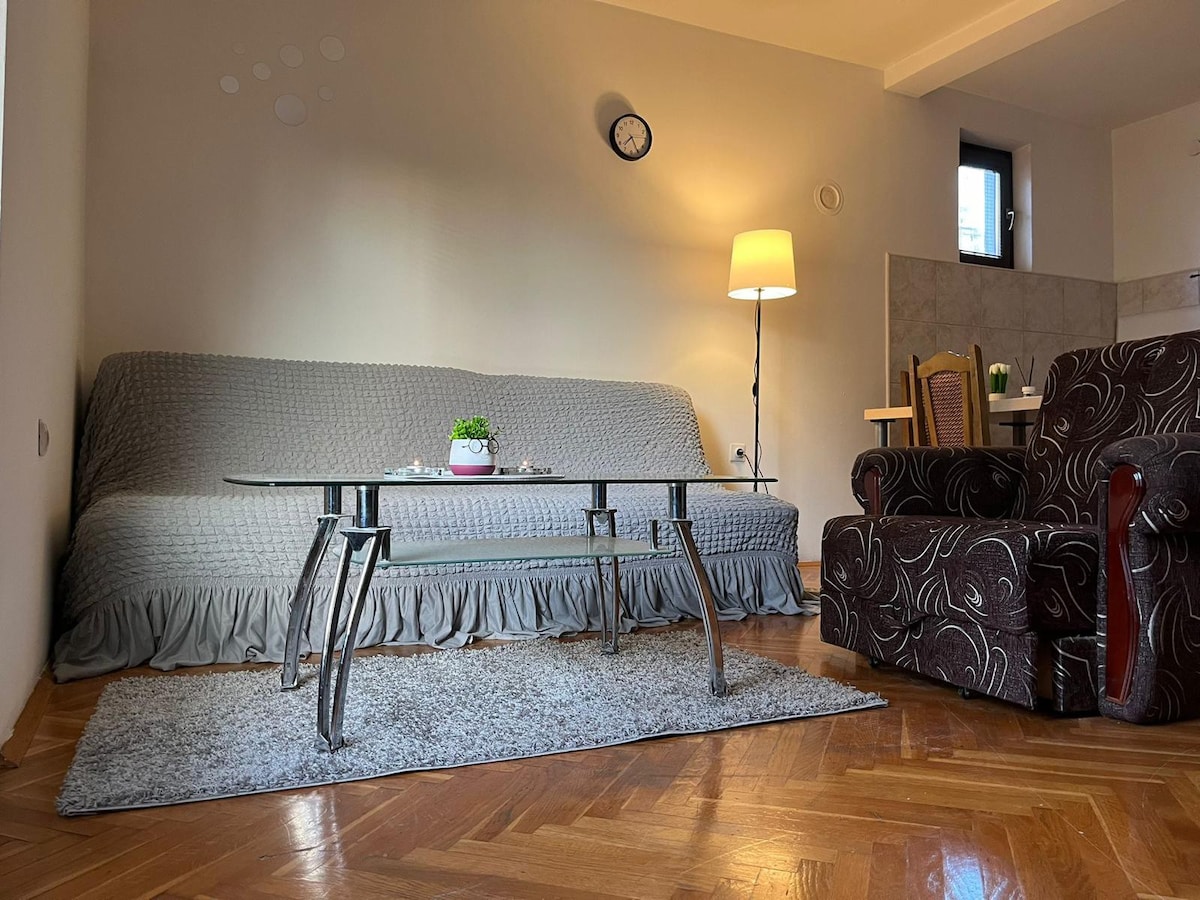 A living area featuring a soft gray sofa covered with a blanket and a glass coffee table. A patterned armchair sits nearby. Natural light enters through a window, illuminating the wooden floor. A small table with two chairs is visible in the background.