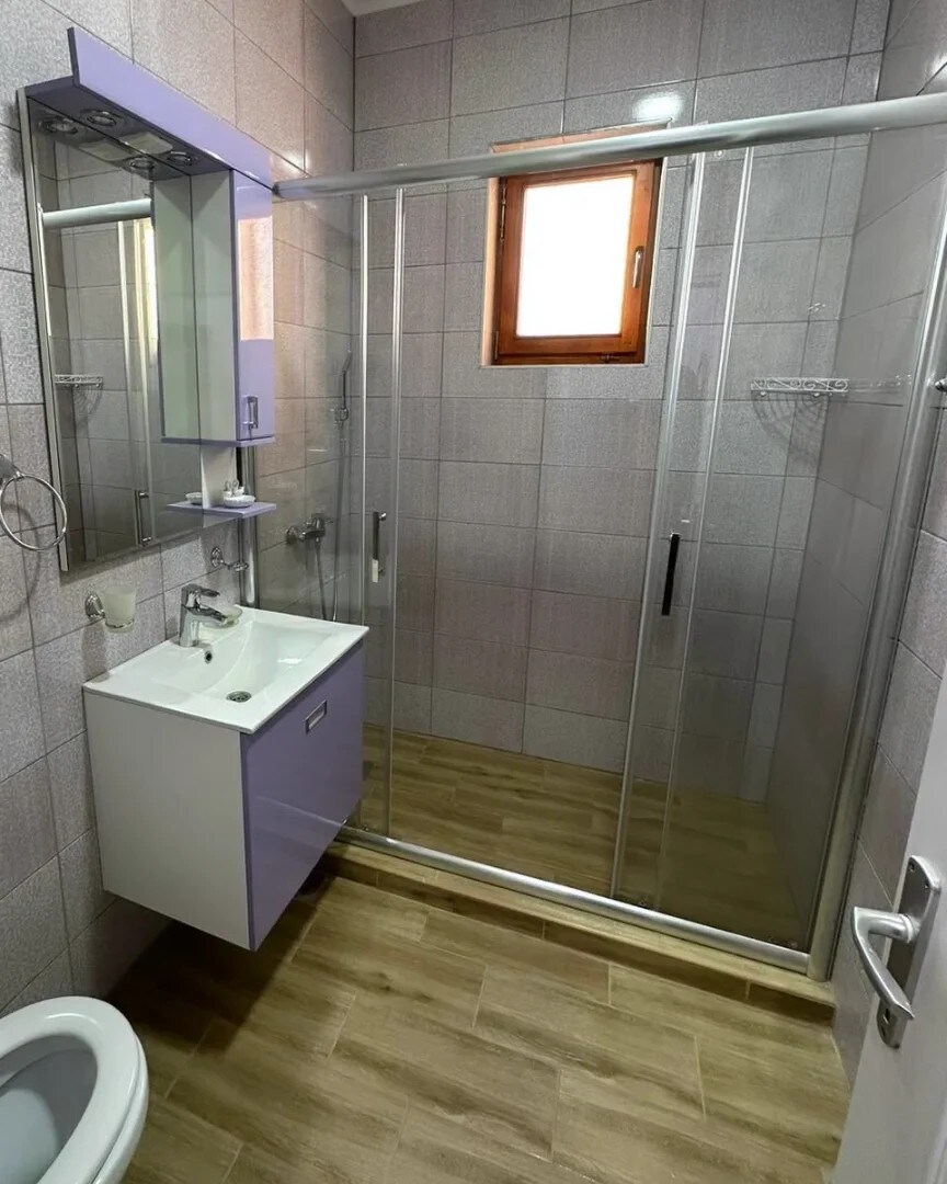 A modern bathroom features a sleek shower enclosure with clear glass doors. A vanity with a minimalist design is complemented by a wall mirror. Natural light enters through a small window, illuminating the tiled walls and floor.