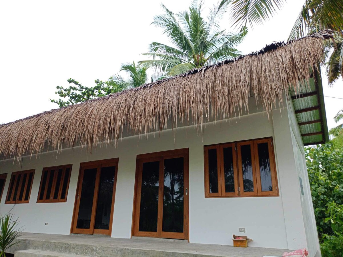 The exterior of a tropical building features a thatched roof and smooth white walls. Large wooden doors and windows, framed in natural wood, reflect the surrounding greenery and palm trees, creating a harmonious connection with nature.